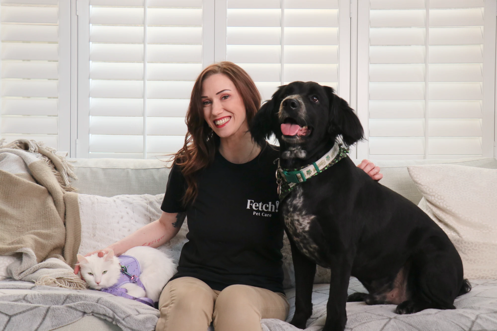 A smiling Fetch! sitter on a couch petting a white cat with a black dog sitting beside her